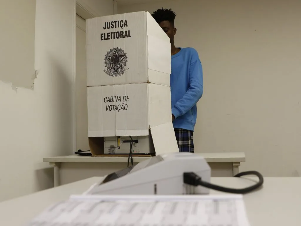 Urna eletrônica em seção eleitoral com policiamento no fundo.